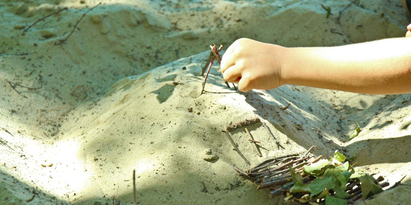 Eine Kinderhand platziert Ästchen und Blätter auf einem kleinen Sandhügel.